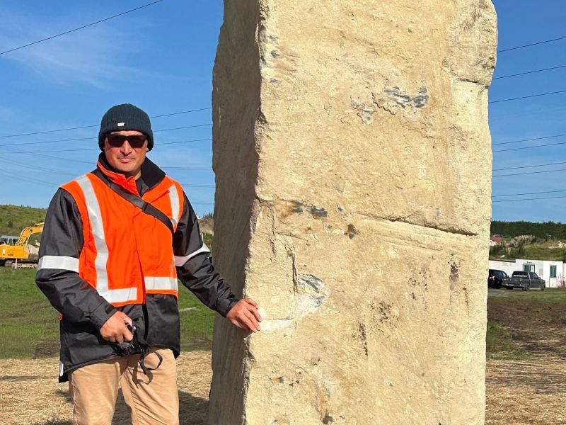 a man in an orange jacket standing next to a large block of sandy coloured stone